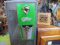 Front view of green Steam Whistle magnetic bottle opener with attached metal cap catcher mounted on a black surface next to wooden furniture and a decorative item.