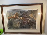 Full view of framed print showing two young cougars resting within rocky terrain and grass.