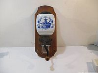 Front view of the ceramic coffee grinder mounted on wood showing the Delft blue windmill design and metal handle.