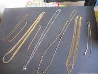 Seven necklaces laid out on a black background, displaying a variety of chain styles from thin delicate chains to thicker braided styles in gold-tone and silver-tone finishes.