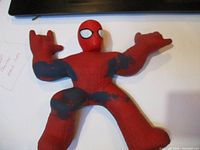 Front view of vintage red and black Spider-Man plush figure with arms and legs spread out on a flat surface. Shows stretched limbs and worn black paint dapples.