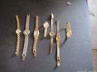 Group shot of seven vintage women's wristwatches in gold and silver tones on a black surface, side view showing bands and faces.