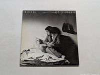Front cover art of Billy Joel's The Stranger LP album, showing Billy Joel sitting next to a masked figure in bed and a shadowy figure holding a hat.