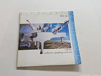 Front cover of the Elton John Live in Australia album showing a white piano and symphony instruments against a white and blue background.