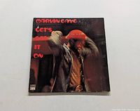 Front cover of Marvin Gaye's Let's Get It On album showing blurred image of artist raising arms wearing a red hat with red text title and artist name.