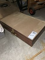 Closed vintage wooden artist case placed on floor, showing exterior with brass handle and latch, rectangular shape 22x13x5 inches.