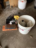 Photo shows a black MotoMaster battery charger with cables, two socket sets in plastic cases (orange and dark blue), and a large white bucket containing bottles of automotive fluids and supplies.