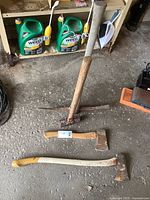 Photo of four vintage tools including a pick axe, axe, hatchet, and sledge hammer on a concrete floor with gardening products in the background.