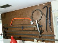 Overview of pegboard with orange frame bow saw, four bar clamps below, two black pry bars on the right