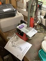 Front view of red vertical belt sander on workbench, showing adjustable aluminum table, sanding belt and attached tag