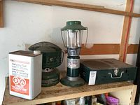 Side view of the near full can of camping fuel, catalytic heater, propane lantern, and the closed Coleman camp stove in metal case.