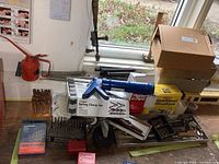 Photo showing assorted hand tools including a box of drill bits, blue caulking gun, spring clamp set in box, red oil can, and other tools on a table near a window.
