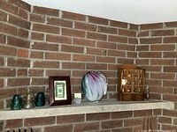 Wide view of mantel showing all items including wooden display case, signed art glass vase, framed Berlin Wall piece with miniature rug, and glass insulators
