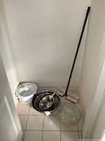 Contents of small closet including Rival crock pot, stainless steel mixing bowls nested together with ladle, a large cut glass punch bowl set with glass cups and lid, and floor duster with black handle and red mop head on tile floor.