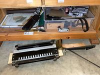 Overall view showing the dovetail jig on the floor beneath a wooden cabinet containing various materials and tools including strap clamps and ratchet straps.