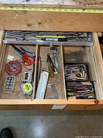 Photo of an open wooden drawer with various hand tools including punches, bits, scissors, tape measures, wrenches, small screwdrivers, and squares arranged in different compartments.