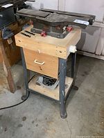 Wide view of entire jointer mounted on metal stand with wooden sawdust catch drawer below, power cord visible on floor