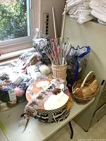 Knitting needles standing in a small woven basket surrounded by yarn bags, round patterned storage box, wicker baskets, and feather headband.