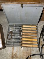 Photo of 7 lathe chisels with wooden handles and 2 additional metal tools in a gray metal case with lid open, showing overall lot presentation and condition.