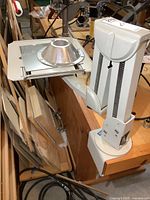 Photo showing the overall heavy-duty clamp-on flexible monitor stand with clamp base attached to a wooden table, flexible arm with springs, and mounting platform with metal plate and cone-shaped adapter.