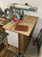 Full view of DeWalt Deluxe Power Shop radial arm saw from front left showing saw blade, arm, and wooden work platform with some dust and wear.