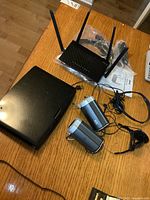 Black Insignia portable DVD player, Asus wireless router with three antennas, two grey Altec Lansing computer speakers, and cables on wooden table.