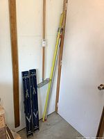 Pair of blue metal folding sawhorses standing folded against a wall next to two yellow poles and a pole sander.