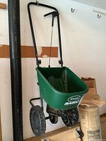 Side and full view of the seed spreader showing black metal frame, handle with lever, and green plastic hopper.