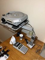 Wide angle showing all items on wooden surface including Kodak slide projector on adjustable clamp base, calculator with paper roll, cordless phone, remote control device, and small container.
