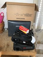 Two Kodak projectors stacked, coiled power cord on top projector, box of bulbs and cardboard margarine box behind