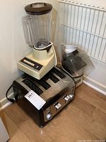 Blender, toaster, and hot air popper grouped together on wooden floor near a white wall and metal rack.