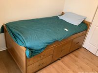 Full view of twin size captain's bed with teal bedding, showing built-in drawers on side and casters on floor.