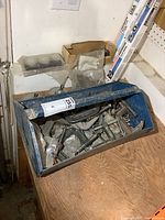 Top-down view of blue metal toolbox filled with C clamps, pipe wrench, and drum sanding pieces on a wooden surface.