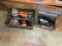Two metal toolboxes on the floor. Left toolbox is dark green with various hand tools. Right toolbox is a silver Porter Cable box containing an electric tool and cord.