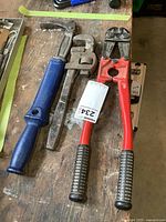 Overview of the three tools laid on the wooden surface showing the blue-handled nail puller, pipe wrench, and red-handled bolt cutters with rubber grips