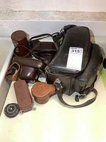 Photo showing black leather camera bag alongside two cameras in brown leather cases, a lens case, and camera accessories spread out on surface.