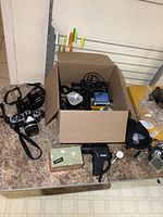 Cardboard box on countertop containing flashgun, cables, battery pack, lens mount ring and small accessories with two cameras flanking box