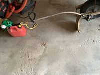 Full view of the weed eater with curved silver shaft, black throttle and handle, yellow pull starter, and red small fuel can. The cutting head has metal guard and line.