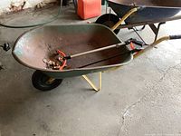 Full side view of the older metal wheelbarrow showing green tub, yellow frame, solid rubber tire, and one grabber tool inside