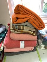 Stack of seven folded wool items: orange, herringbone, plaid, peach, coral, pink, grey