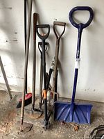 Photo of a collection of yard tools against a wall showing an edger, hoe, fork, snow shovel, weed puller, and other tools in used condition.