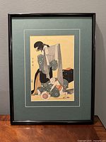 Framed Japanese woodcut print showing Geisha and child, displayed against grey wall