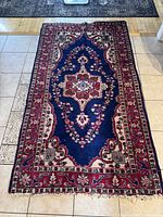 Full view of the vintage Persian wool area rug laid out on a tile floor, showing navy blue central field with red floral border and intricate medallion