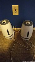 Two white Sunbeam humidifiers with gray accents side by side on carpet in front of a blue wall with power outlet.