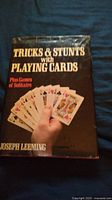Front cover photo of 'Tricks & Stunts with Playing Cards' book showing title and hand holding playing cards with some wear on dust jacket.