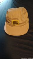 Khaki five-panel Good Supply hat on dark surface showing front and side detail.