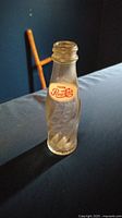 Photo of small vintage Pepsi Cola glass bottle upright on a dark surface, showing embossed twisted pattern and red and white label