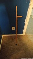Full view of the maple wood walking cane standing against a blue wall and carpet floor, showing its straight shaft, side handle, and rubber tip.