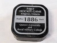 Top side of puck container showing text for first hockey game replica puck - 1886, Queen's University vs Royal Military College