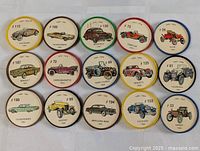 Lot of 16 vintage Jell-O car coins, each featuring illustrations of classic cars with labels including make, model, and year, arranged in rows on a white fabric background.
